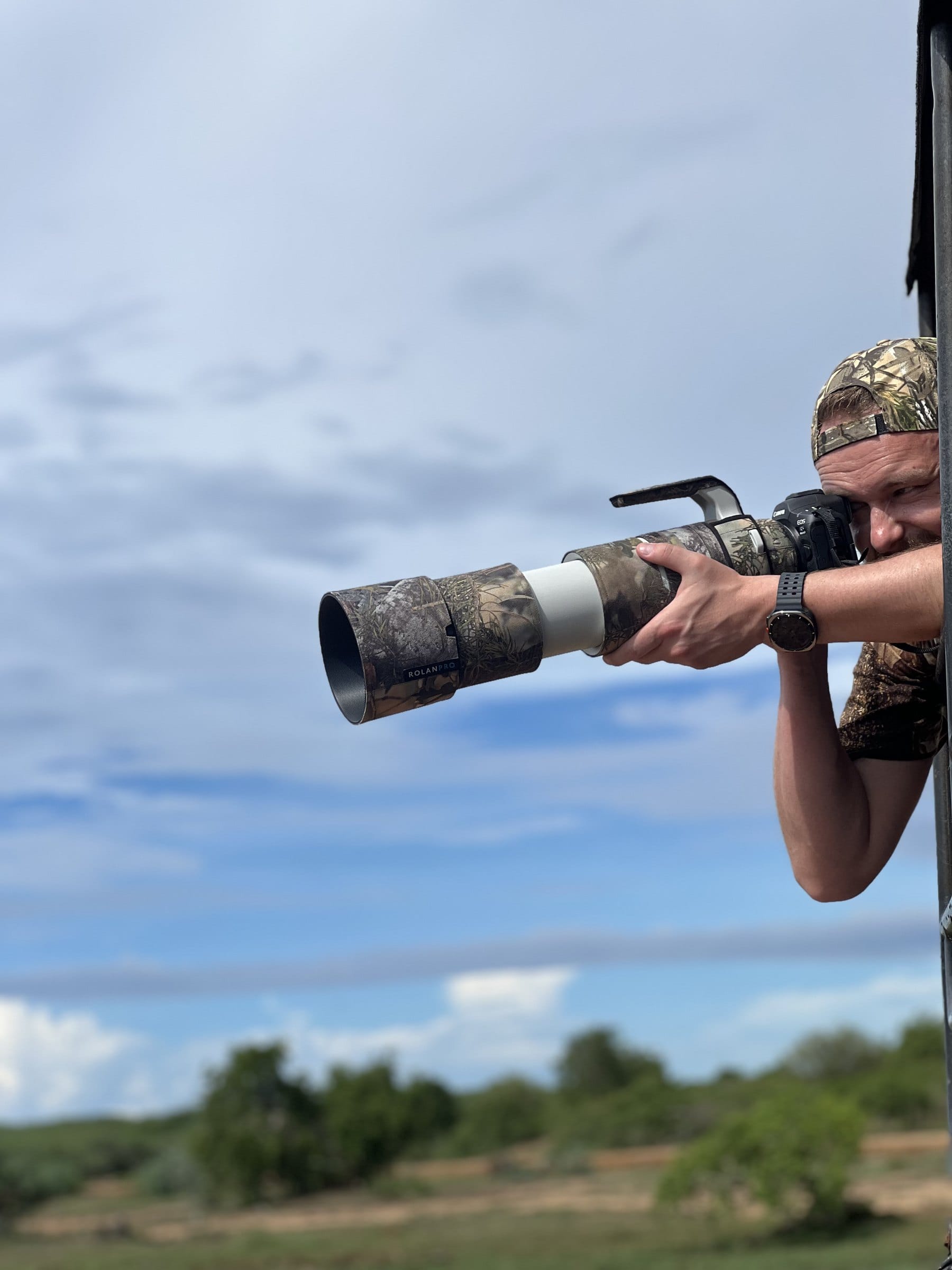 A wildlife photographer leaning out of a safari jeep with a camouflaged telephoto lens
